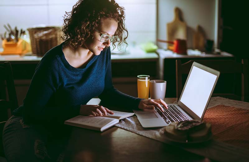 woman paging through notes sitting on laptop at kitchen table