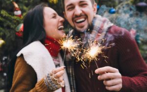 A couple holding sparklers in winter attire in front of a Christmas tree smiling and enjoying the space.
