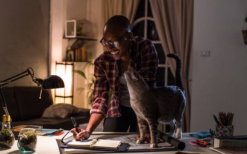 Person writing in a notebook while a cat rubs their face
