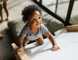 Smiling toddler with curly hair crawling on white bedding in bright nursery